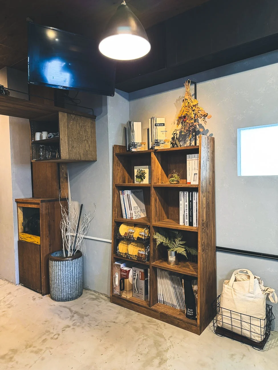 木製の書棚を中心に、本や雑誌、植物、ドライフラワーなどが飾られたおしゃれなインテリア空間です。壁掛けテレビやペンダントライトも配置され、落ち着いたカフェのような雰囲気が漂っています。