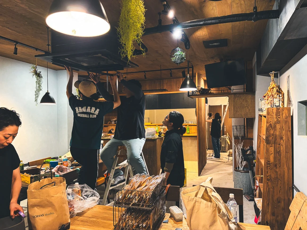 カフェのような店内で数人の男女が作業をしている様子。2人の男性が脚立を使って天井の照明器具を設置しており、他のスタッフも準備を進めている。開店準備や改装中の活気ある店内風景です。