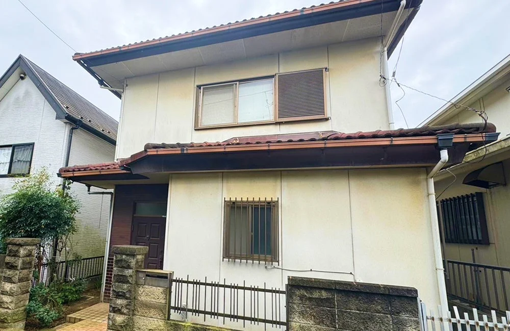 曇り空の下、やや老朽化が見られる日本の二階建て住宅の外観です。茶色の瓦屋根と雨樋が特徴で、玄関前には石造りの門柱とフェンス、植栽が見えます。