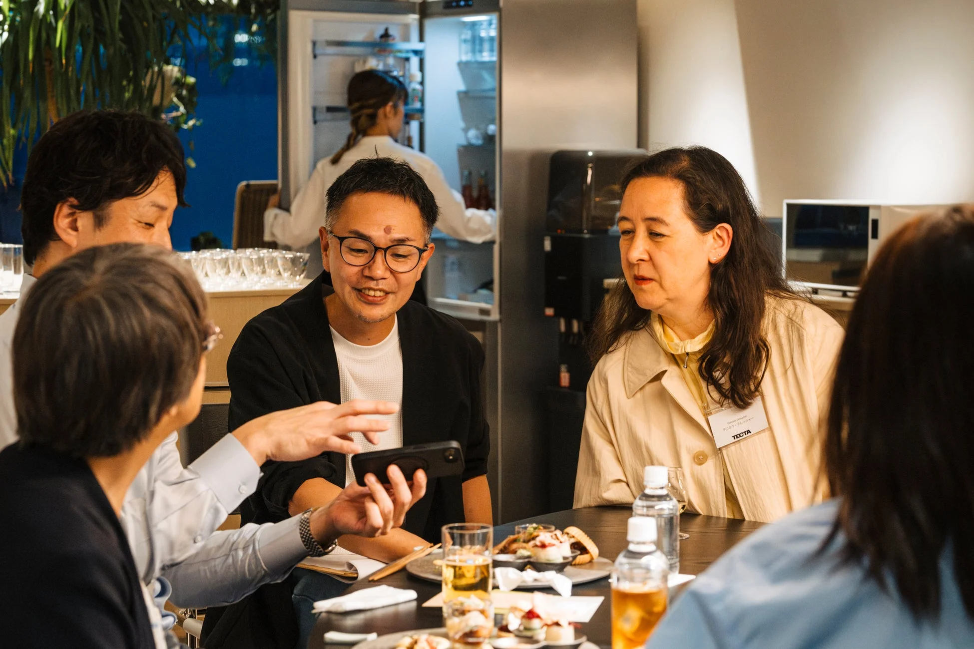 カジュアルな雰囲気の中で、数人がテーブルを囲んで交流している様子。一人がスマートフォンを手に何かを見せ、他のメンバーは話に耳を傾けている。