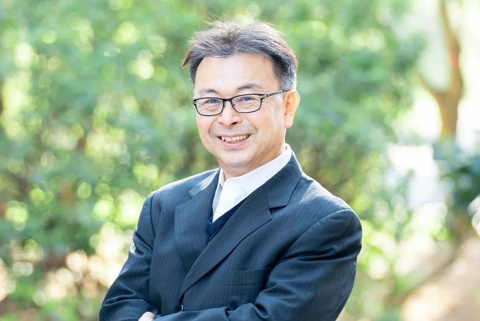緑の背景を背に、スーツと眼鏡を着用した中年の男性が、明るく親しみやすい笑顔でカメラを見つめているポートレート写真です。