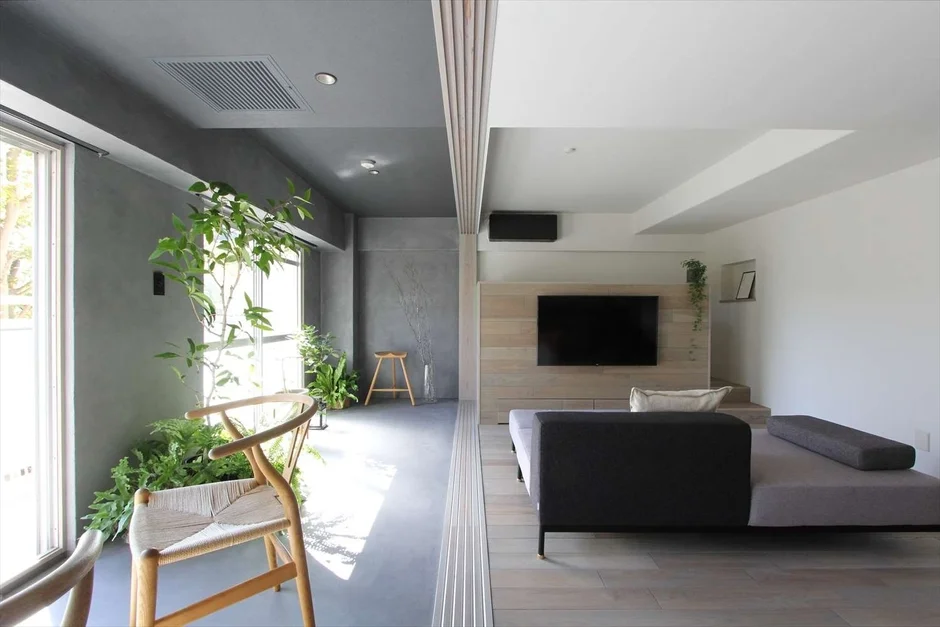 大きな窓から光が差し込むモダンなリビングルーム。観葉植物が豊かな空間と、ウッドパネルの壁にテレビが配された落ち着いたエリアが融合しています。コンクリートと木の素材感が調和した、洗練されたデザインの住空間です。