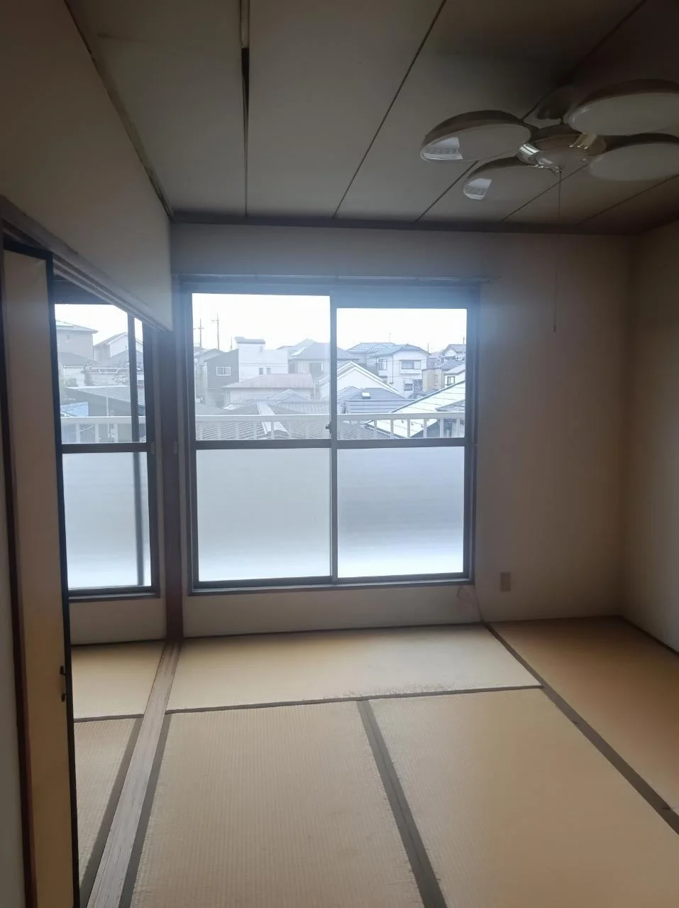 畳敷きの和室で、窓からは住宅街の風景が見えます。天井には照明があり、部屋は空室で家具はありません。年季の入った内装で、リフォーム前の状態のようです。