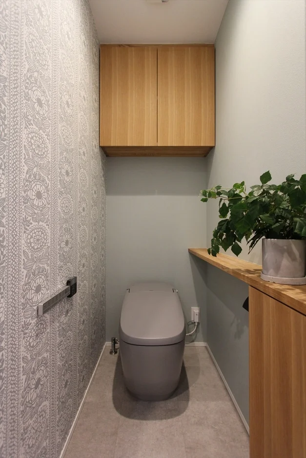 グレーのタンクレストイレが設置された、モダンで清潔感のあるトイレ空間です。左側の壁には柄物のアクセントクロスが施され、上部には木目調の収納棚があります。右側の棚には観葉植物が置かれ、落ち着いた雰囲気を演出しています。
