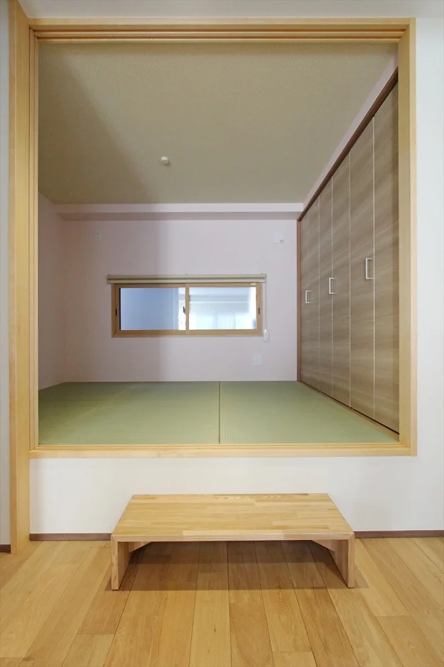 フローリングの部屋から続く小上がりの和室のインテリアです。畳が敷かれた和室には窓と大型クローゼットがあり、手前には木製のベンチが置かれ、落ち着いた空間を演出しています。