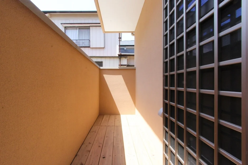 ベージュ色の壁と木製の床が特徴的な、日差しが差し込む狭いバルコニーの風景です。床には斜めに影が落ちており、右側には格子状の構造物が見えます。背景には他の建物が確認できます。