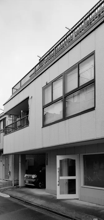白黒で撮影された日本の都市部の建物。1階部分はピロティ式の駐車場になっており、黒い車が停まっている。白い扉がわずかに開いており、上階には窓やバルコニーが見える。