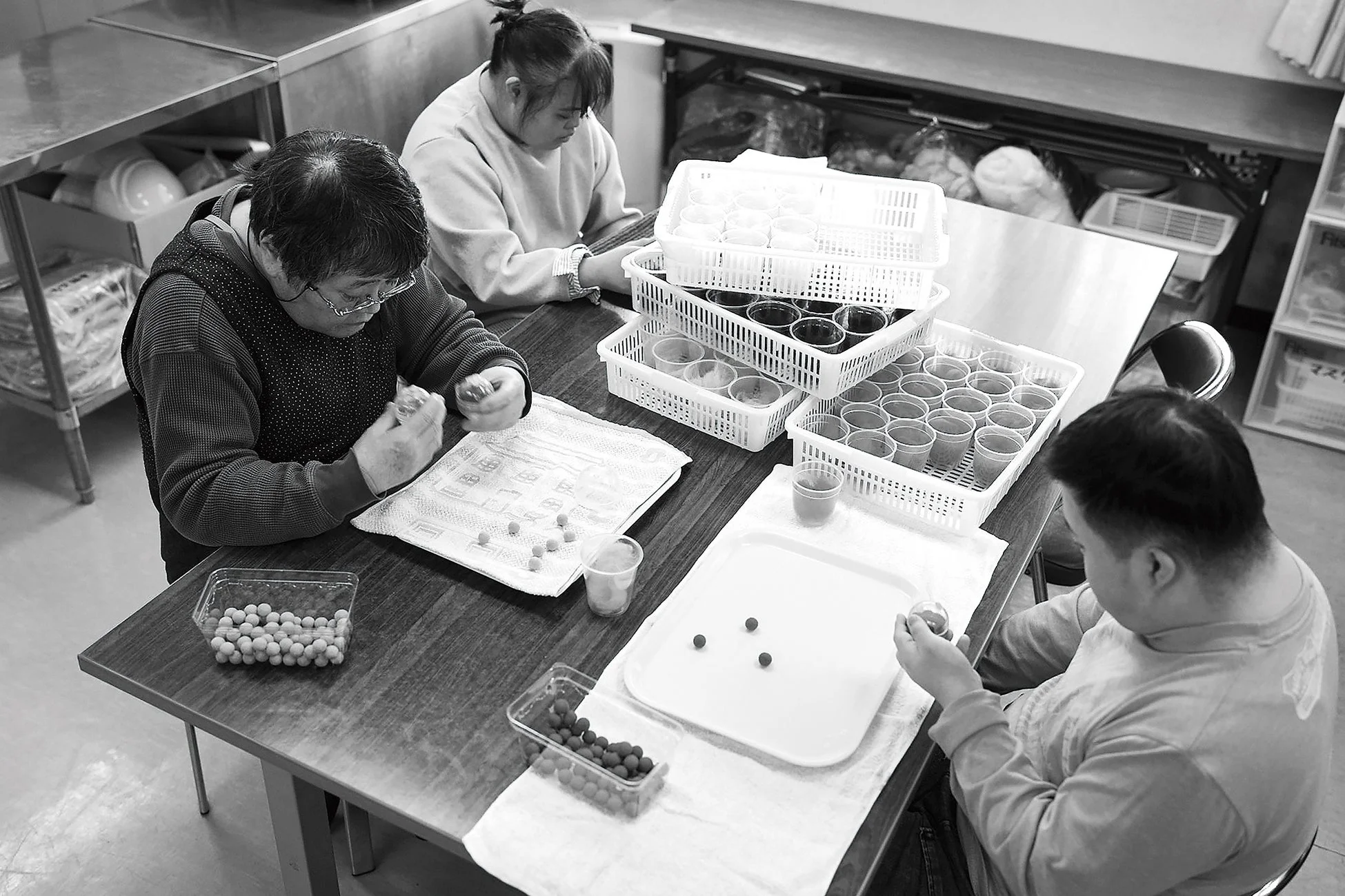 3人がテーブルを囲んで座り、小さな物体を扱って集中して作業している様子です。白いカゴに入った多数のカップや、丸い粒状のものが置かれており、福祉施設や作業所での軽作業や訓練の一環と思われます。