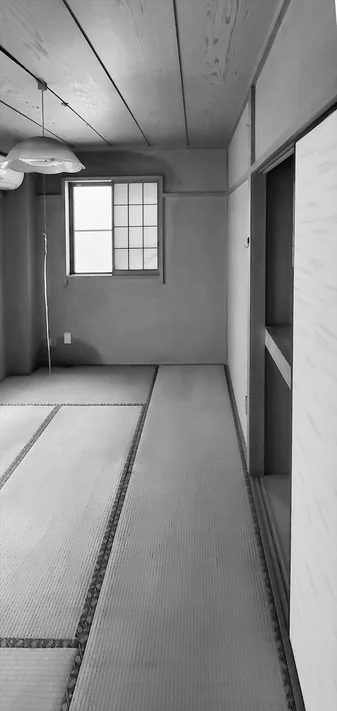 畳が敷かれた和室の内部を白黒で撮影した写真。窓や木目の天井、天井照明が見える。静かで unoccupiedな空間の雰囲気を伝えています。