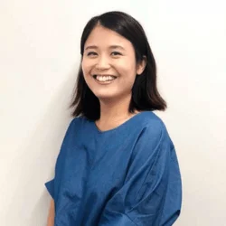 白い背景の前で、青い服を着た短髪のアジア人女性が笑顔で正面を向いている上半身のポートレート写真です。