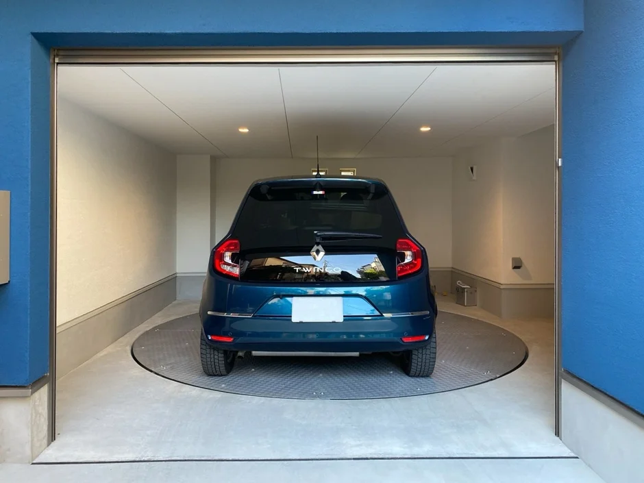 青いルノー・トゥインゴが、車を回転させるためのターニングプレートを備えたモダンなガレージ内に駐車されています。ガレージの壁は白く、入り口のフレームは鮮やかな青色です。