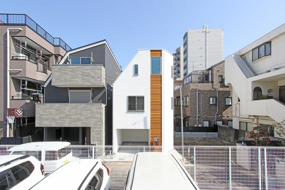青空の下、密集した住宅街に建つ、細長い白い現代的な住宅