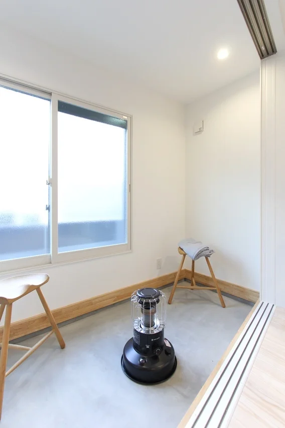 白を基調としたシンプルな部屋に、窓、木製スツール、そして「Snow Peak」の黒い灯油ストーブが置かれている