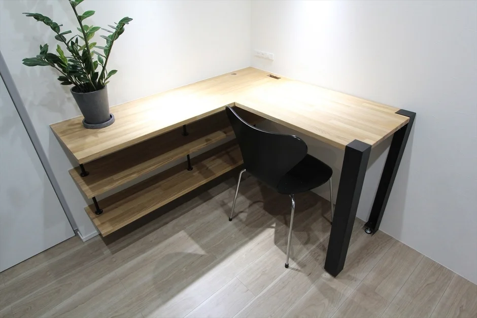 白い壁と木目のフローリングの部屋に、L字型の木製デスクと黒い椅子が配置されています。デスクの棚には観葉植物が置かれ、シンプルでモダンなワークスペースを構成しています。