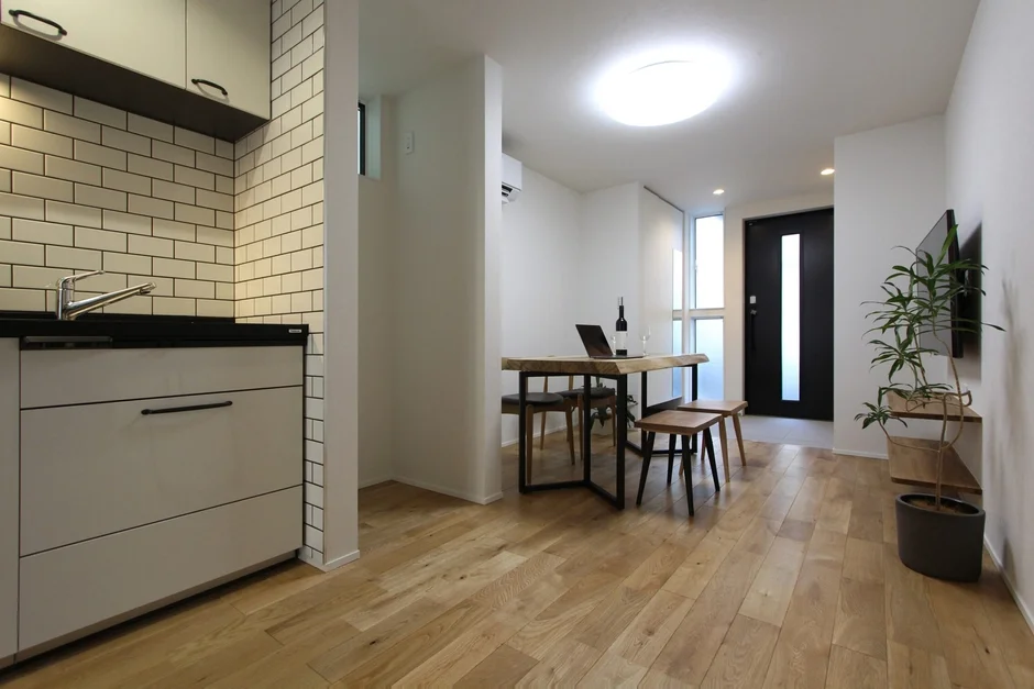 明るくモダンなワンルームマンションの室内。コンパクトなキッチン、木製のダイニングテーブルにはノートパソコンとワインが置かれ、観葉植物が配置されている。白を基調とした壁と木目の床が清潔感のある空間を演出しています。