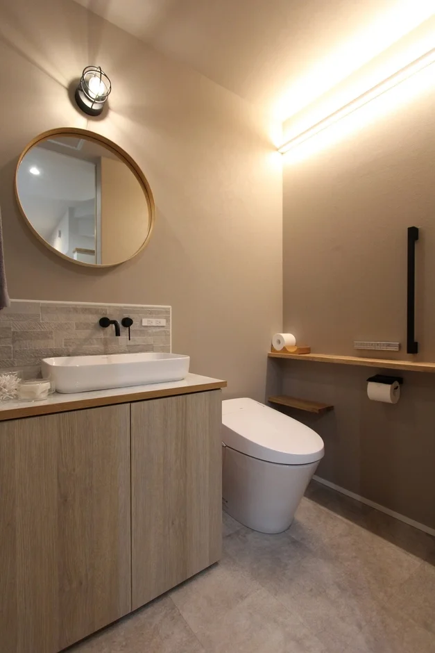 ベージュの壁と木目調の洗面台が温かい雰囲気の、モダンで清潔感のあるトイレ兼洗面空間です。丸い鏡と間接照明がスタイリッシュな印象を与え、機能的な収納も備わっています。