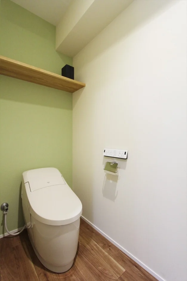 明るい木目のフローリングと、淡い緑と白のツートンカラーの壁が特徴的なモダンなトイレ空間です。上部には木製の棚が設けられ、機能的なタンクレストイレが設置されています。
