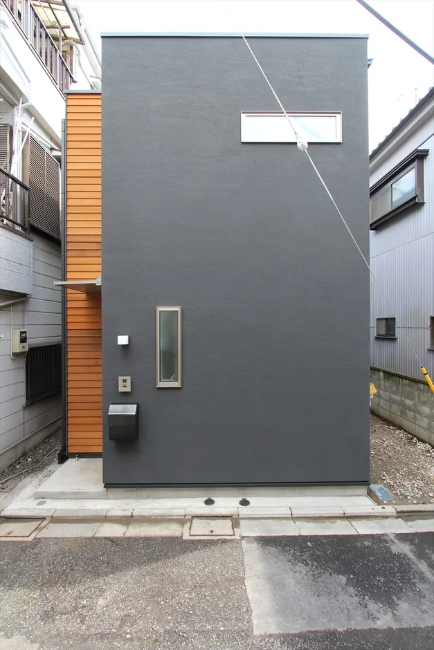 都市部に建つ、グレーのシンプルな外壁と木材のアクセントが特徴的な現代的な狭小住宅のファサードです。