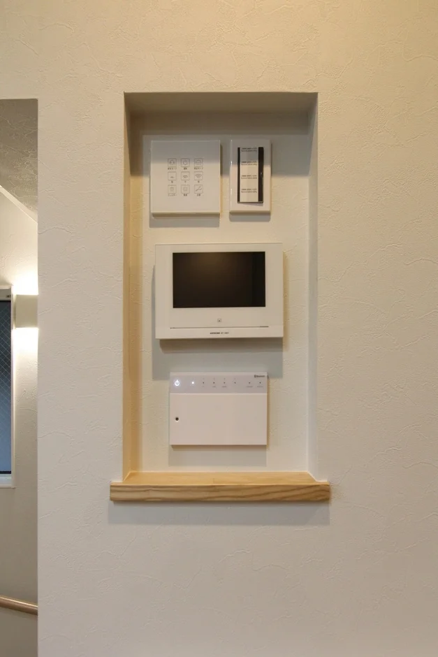 白い壁のニッチに、スマートホームの操作パネル、ビデオインターホン、その他の住宅設備が埋め込まれています。下部には木製の棚があり、機能的で整頓された印象を与える壁面です。