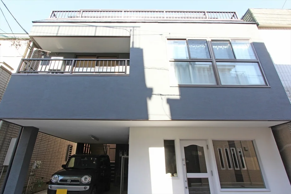 グレーと白を基調としたモダンな3階建て住宅の外観写真です。2階にはバルコニーがあり、1階部分はピロティ形式の駐車場になっており、黒い車が停まっています。日差しが強く、建物の影が印象的です。