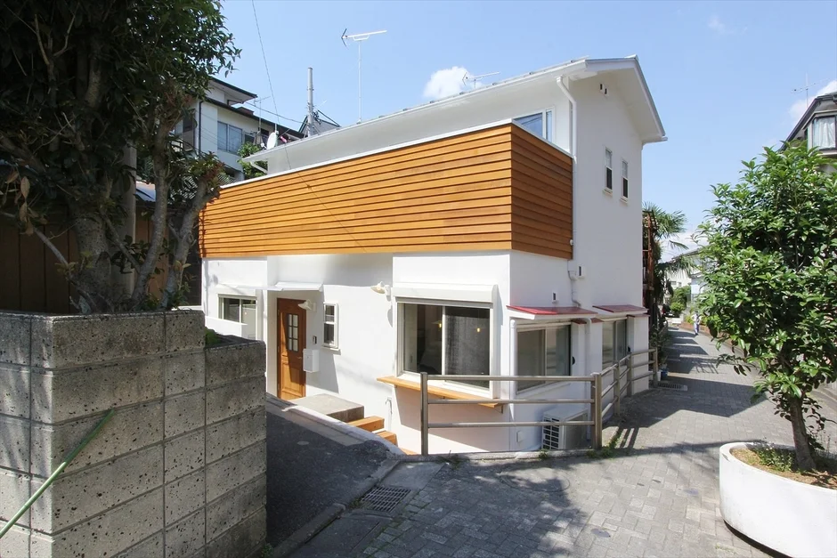 白い外壁と木目調のアクセントが特徴のモダンな一軒家が、青空の下、坂道の住宅街に建っている様子を捉えた画像です。