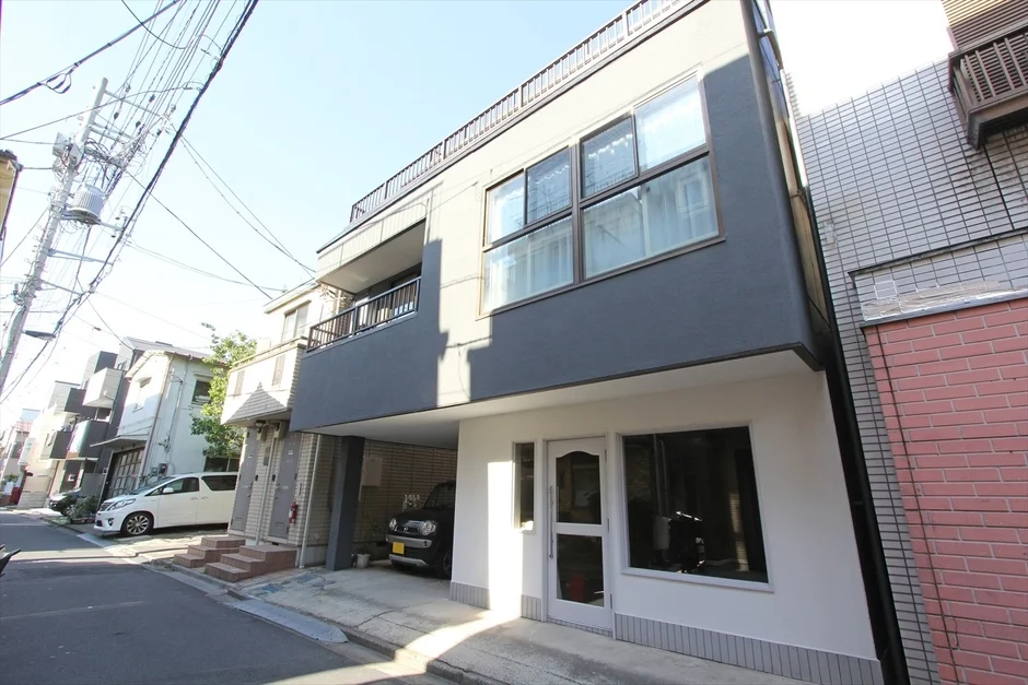 青空の下、モダンなグレーと白の外観が特徴的な建物が写っています。1階は店舗または入口、上階は住居で、建物下には駐車場があります。電線や電柱が目立つ日本の住宅街の風景です。