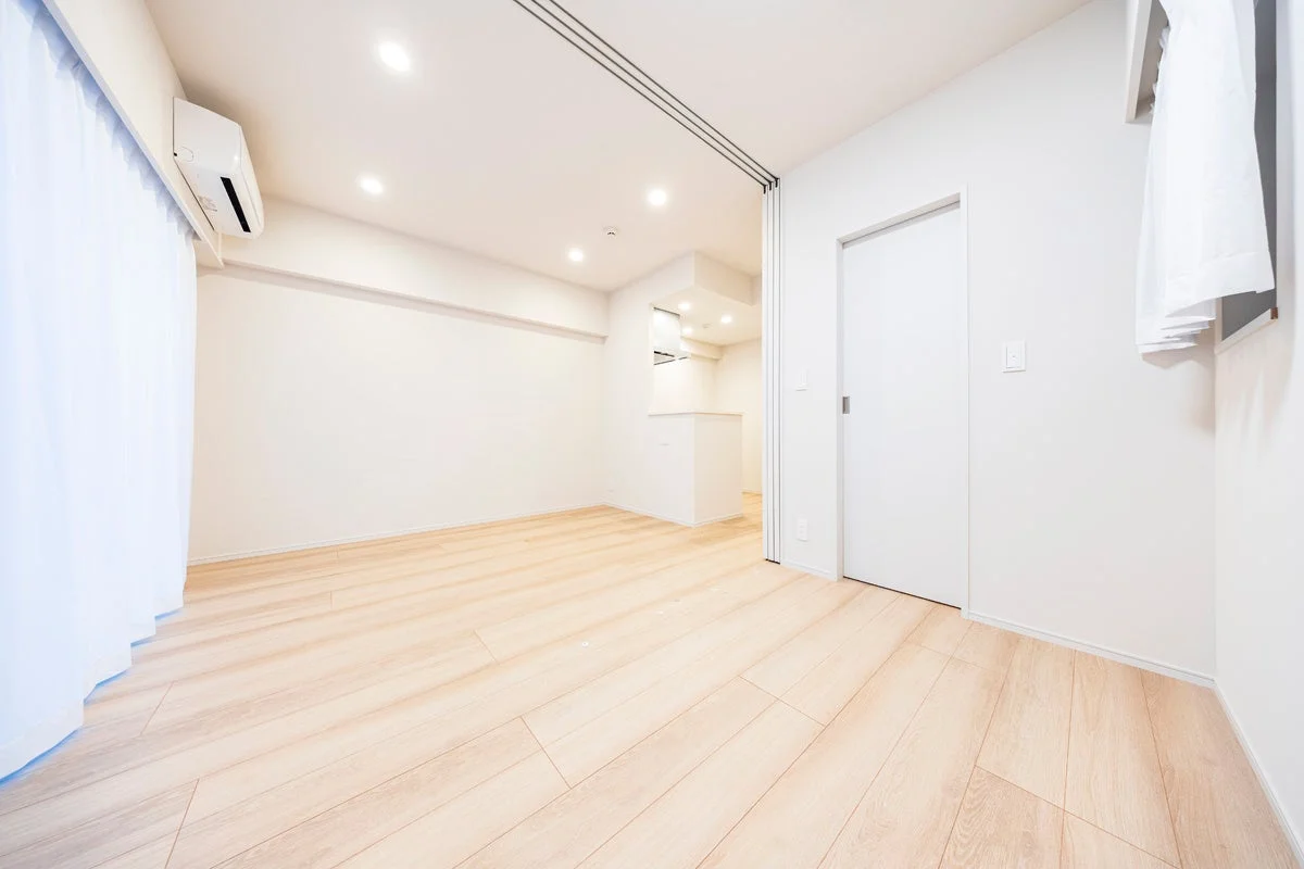 白を基調とした明るく清潔な空室の部屋。フローリングの床、エアコン、ダウンライトが設置されており、奥にはキッチンカウンターが見える。モダンでシンプルな内装が特徴