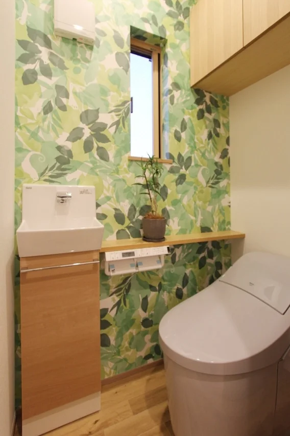緑の葉っぱ柄の壁紙が目を引く、明るく爽やかな印象のトイレ空間です。モダンなデザインの便器と手洗い器が設置され、小窓や観葉植物が心地よい雰囲気を作り出しています。