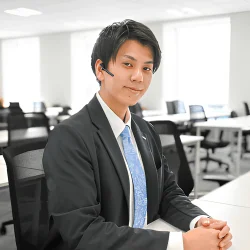 オフィスでスーツを着用した若い男性がデスクに座り、ヘッドセットを付けてカメラを見ています。ビジネスシーンにおける真面目な雰囲気が伝わる一枚です。