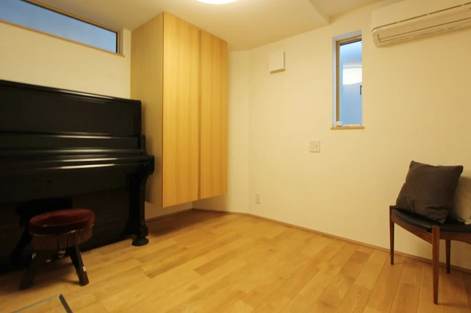 明るいフローリングの部屋に、黒いアップライトピアノと木製の吊り戸棚が配置されています。窓、エアコン、椅子も見えるシンプルで機能的な空間です。