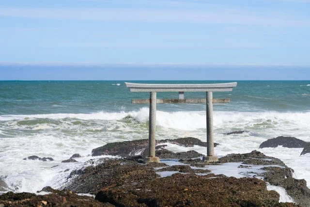 荒々しい波が打ち寄せる岩場に立つ石造りの鳥居が特徴的な海の風景です。晴れた日の青い空と広がる海が印象的です。