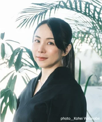 自然光が差し込む場所で、黒い服を着たアジア人女性のポートレート。彼女はカメラに向かって穏やかに微笑み、背景には緑の植物が見える。長髪をポニーテールにまとめ、真珠のネックレスを着用している。