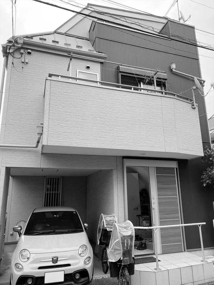 モノクロで撮影された日本の住宅地の建物です。1階部分にはガレージがあり、白い小型車が駐車されています。
