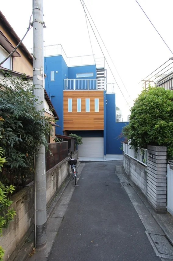狭い路地の奥に立つ、鮮やかな青と木製パネルが特徴的なモダンな住宅。電柱や植栽、自転車が日本の住宅街の日常風景を構成している。