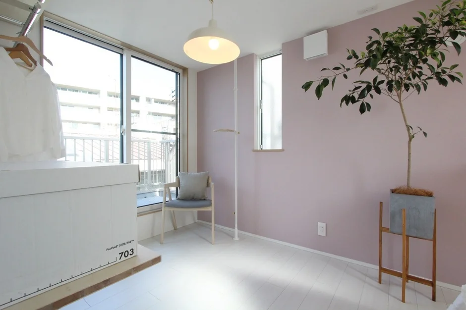 明るい自然光が差し込む、ピンクの壁と白いフローリングが特徴的なシンプルな部屋。観葉植物、白い椅子、照明が配置され、窓からは外の景色が見える。収納ボックスやハンガーラックもあり、機能的で落ち着いた空間です。