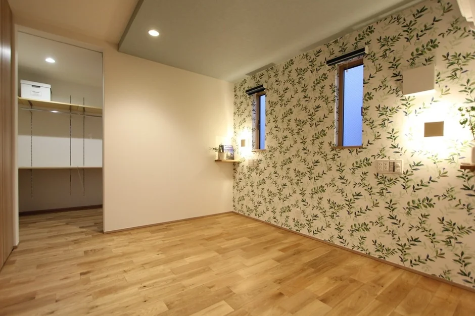 明るい木目調のフローリングと、緑の葉っぱ柄の壁紙が特徴的な部屋です。壁には小さな窓と間接照明があり、開放的なクローゼットも備わっています。清潔感があり、様々な用途に使えるモダンな空間です。