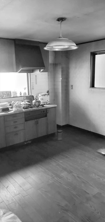 白黒で撮影された生活感のあるキッチンの様子です。調理台には鍋や食器、炊飯器などが置かれ、上部にはレンジフードとペンダントライトが見えます。床は木目調です。