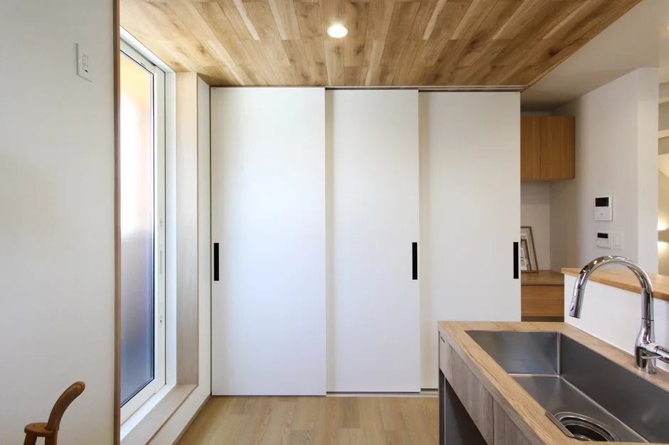 木目調の天井とフローリングが特徴的なモダンな住宅の室内空間です。キッチンカウンターにはシンクがあり、奥には白い引き戸が見えます。
