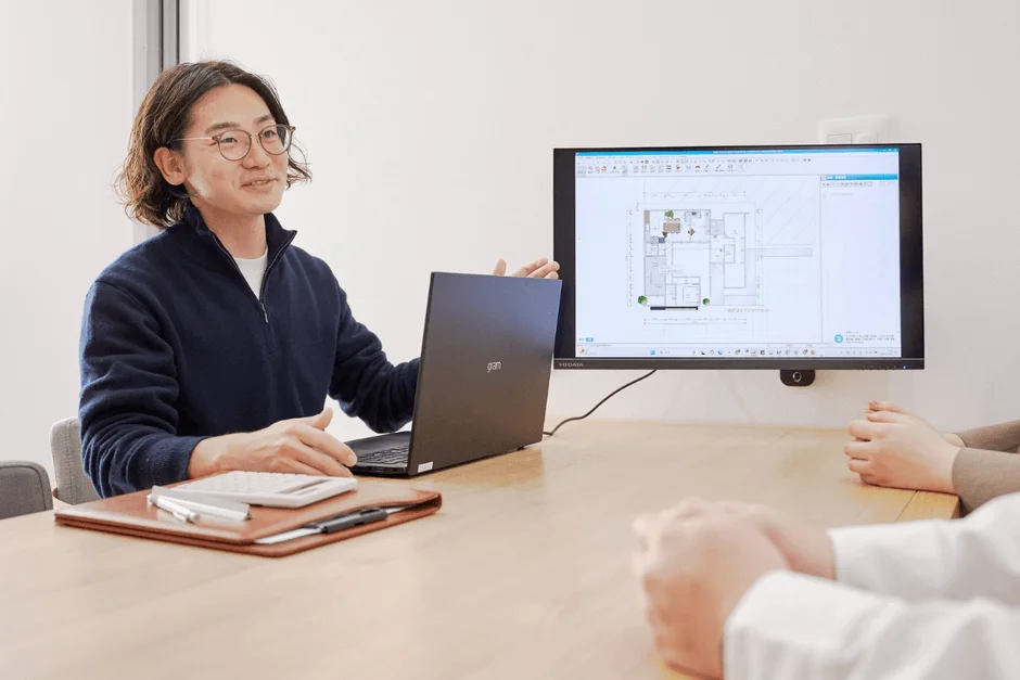 オフィスで男性がノートパソコンとモニターに表示された建築設計図を説明している。会議室での打ち合わせやコンサルティングの様子がうかがえる。