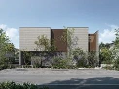 青空の下、道路に面して建つモダンでシンプルなデザインの住宅の外観です。建物はベージュとブラウンのツートンカラーで、前面には植栽が豊かに配置されています。