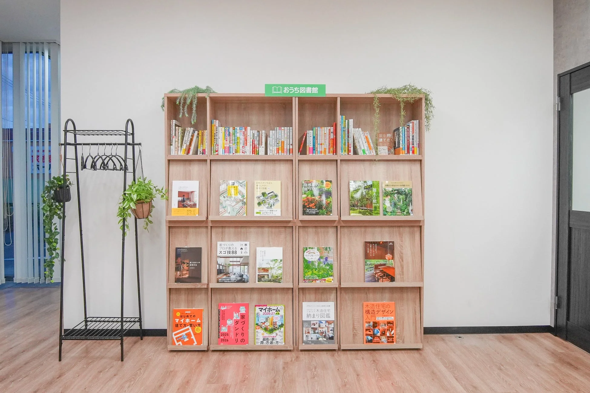 「おうち図書館」と書かれたサインがある木製の大きな本棚が特徴的な、明るくモダンな空間です。本棚には住宅、ガーデニング、ライフスタイルに関する書籍が並び、隣にはハンギングプランターが飾られた黒いスタンドがあり、緑が空間に彩りを添えています。