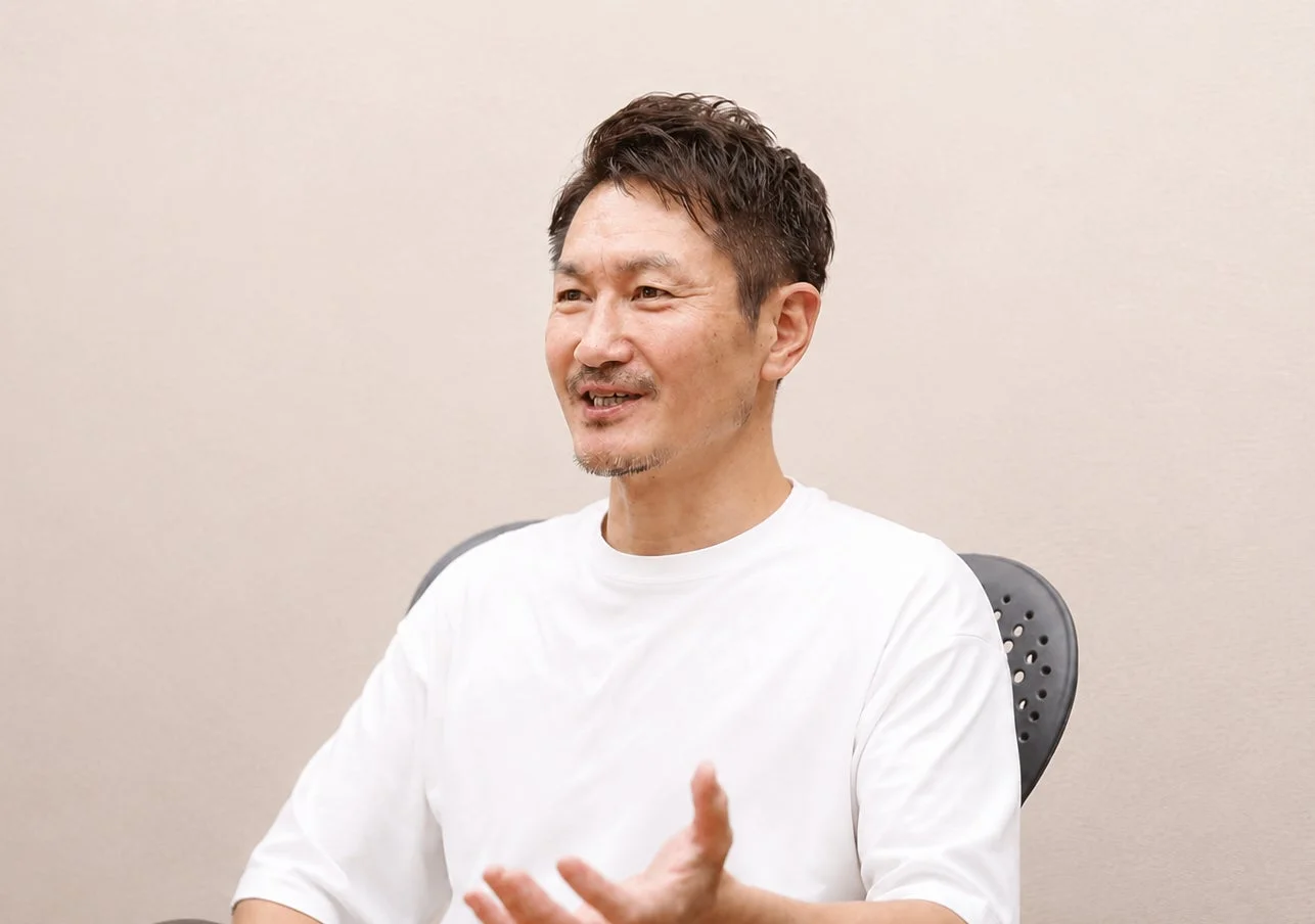 白いTシャツを着たアジア人男性が、笑顔で何かを話しながらジェスチャーをしている様子を捉えたポートレート写真です。