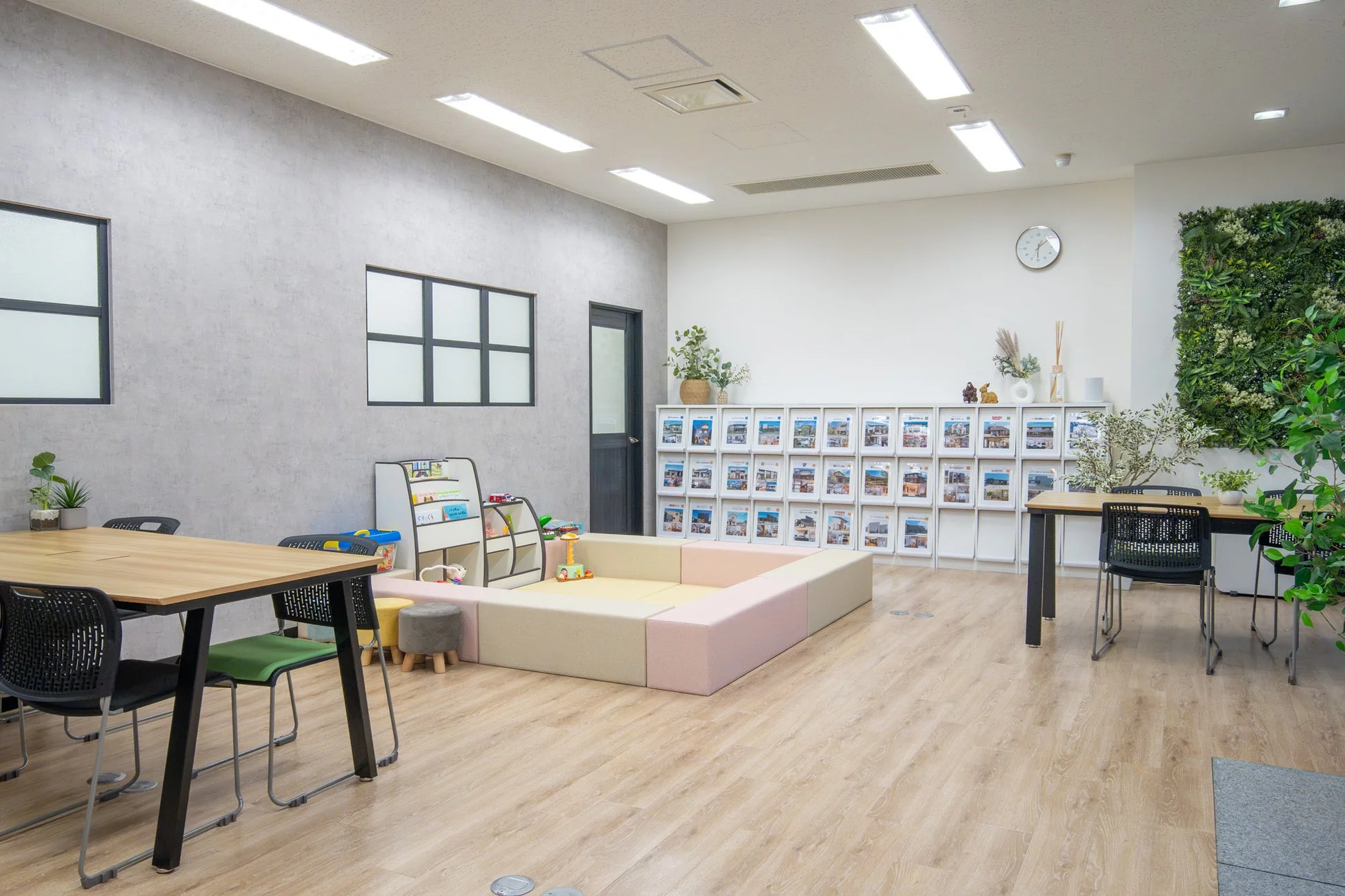 モダンで明るい雰囲気の多目的スペース。商談や休憩に使えるテーブル席、子供が遊べるキッズスペース、住宅情報などのパンフレットが並ぶ棚、そして壁面緑化が配置されており、広々とした清潔感のある空間です。